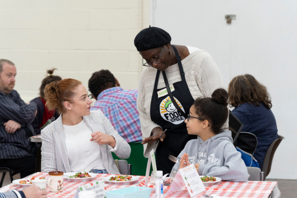 Child and Adult eating a community meal with volunteer speaking to them.